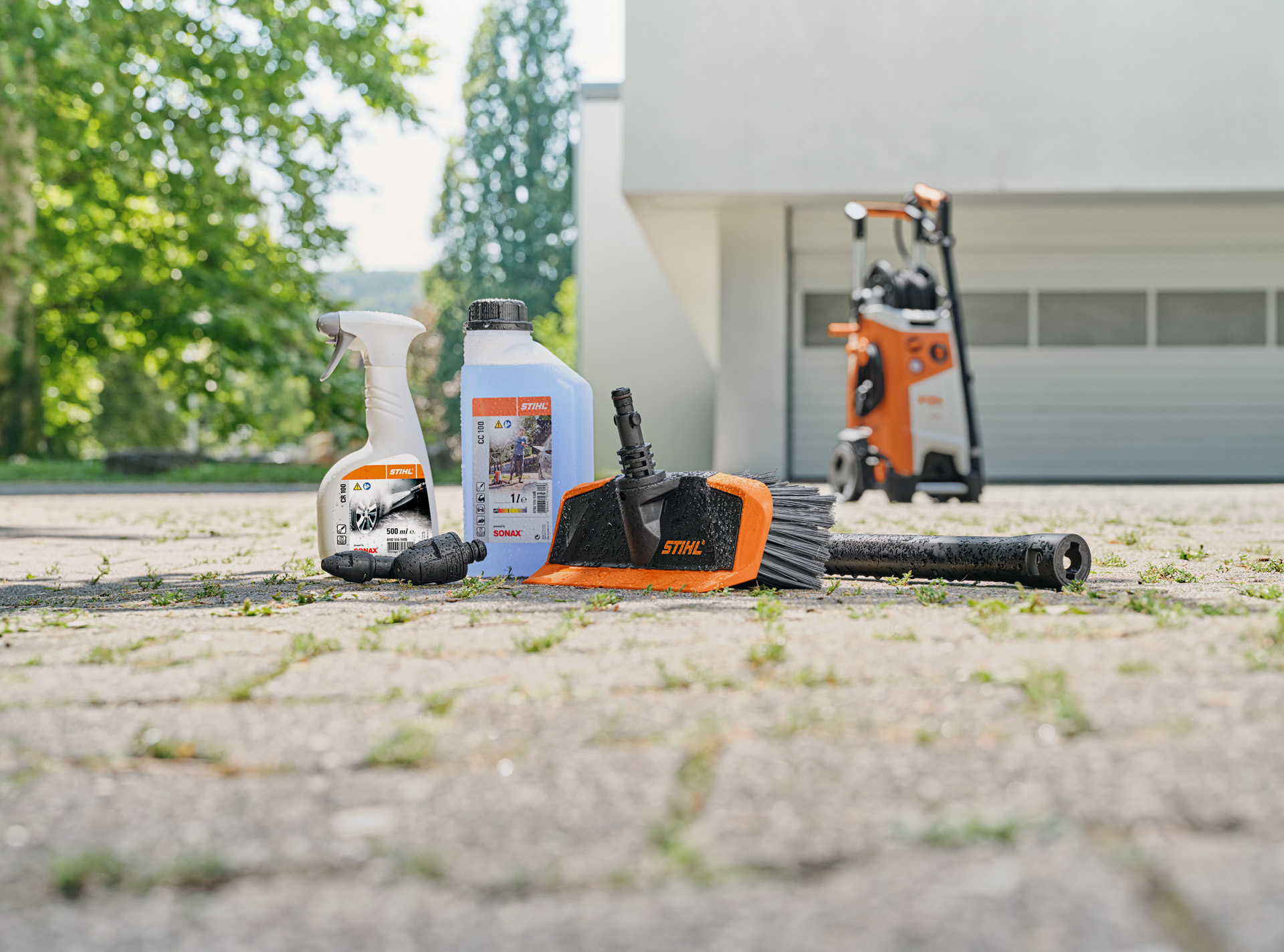  STIHL sredstvo za čišćenje naplataka, šampon za pranje vozila i četka za pranje na tlu ispred bele garaže. U pozadini je STIHL uređaj za pranje pod visokim pritiskom